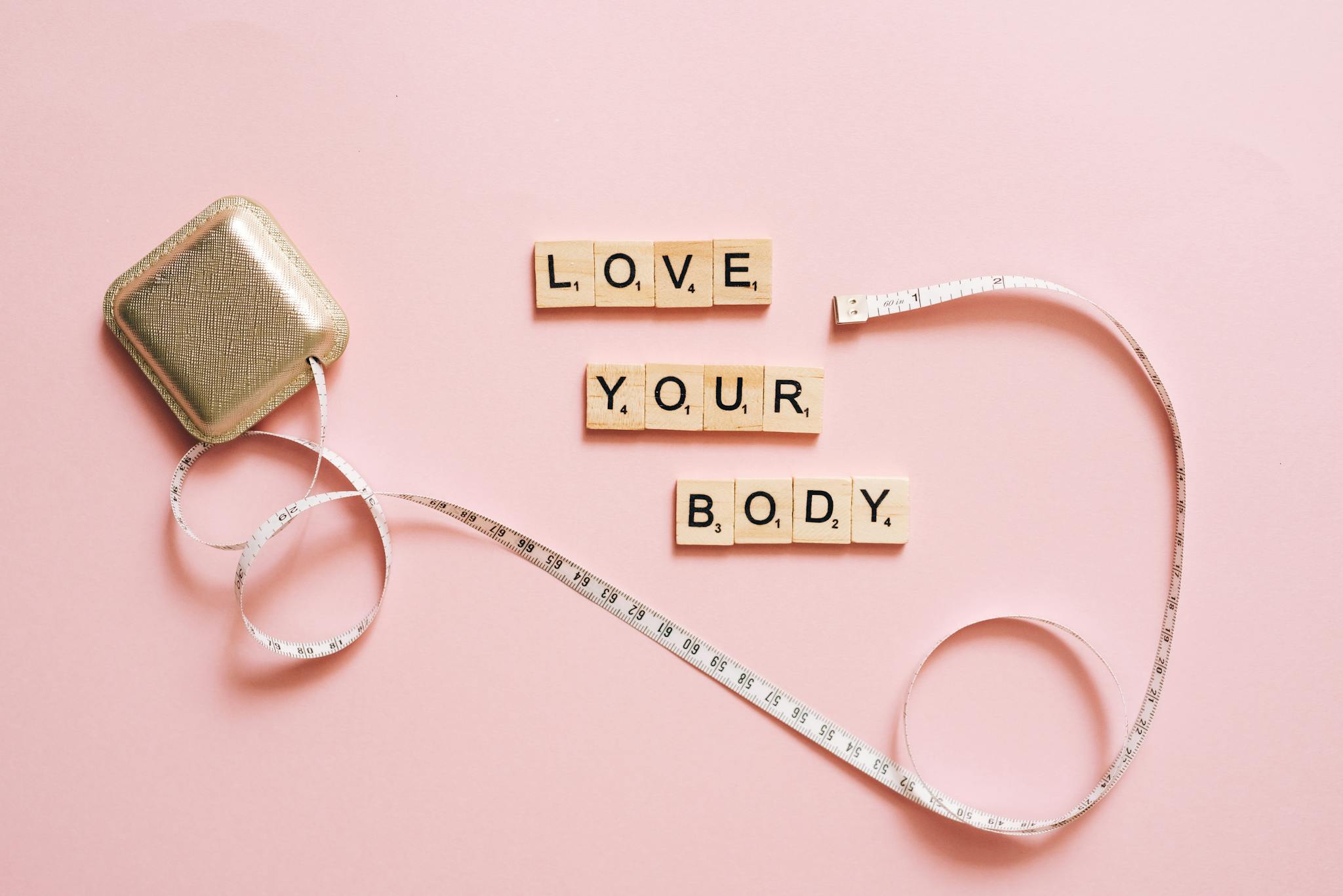 Scrabble letters spelling 'Love Your Body' with a tape measure on a pink backdrop.